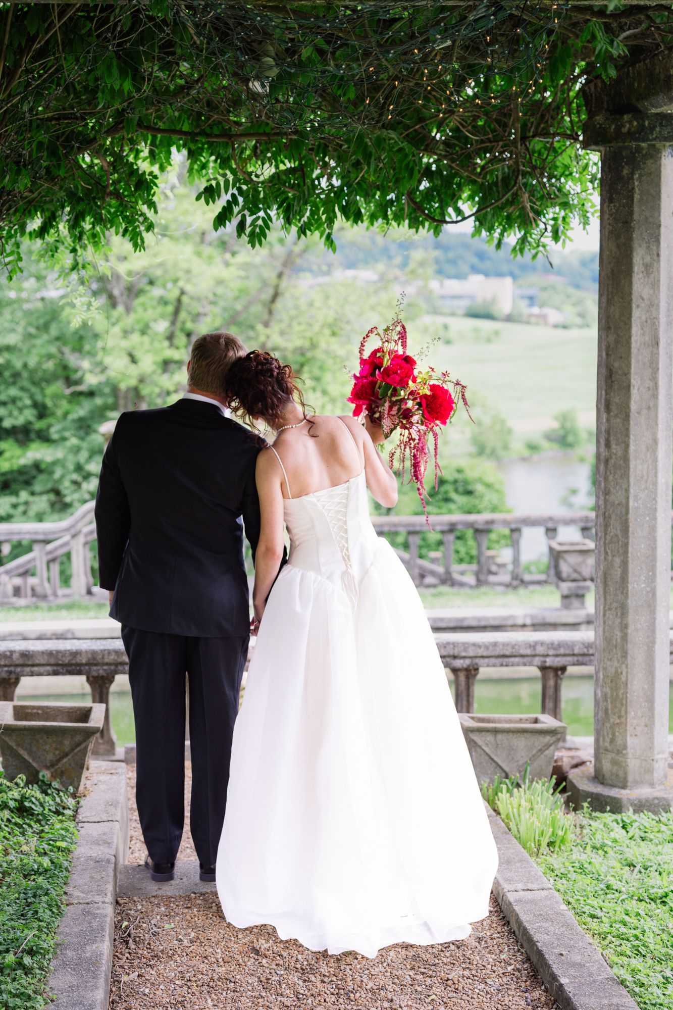A couple in wedding attire walk together under a columned pergola surrounded by lush greenery in a garden setting.