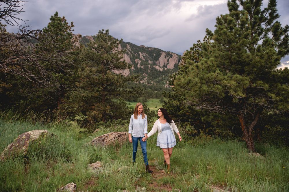 Two people walking through a grassy mountain meadow with pine trees and rocky cliffs in the background.