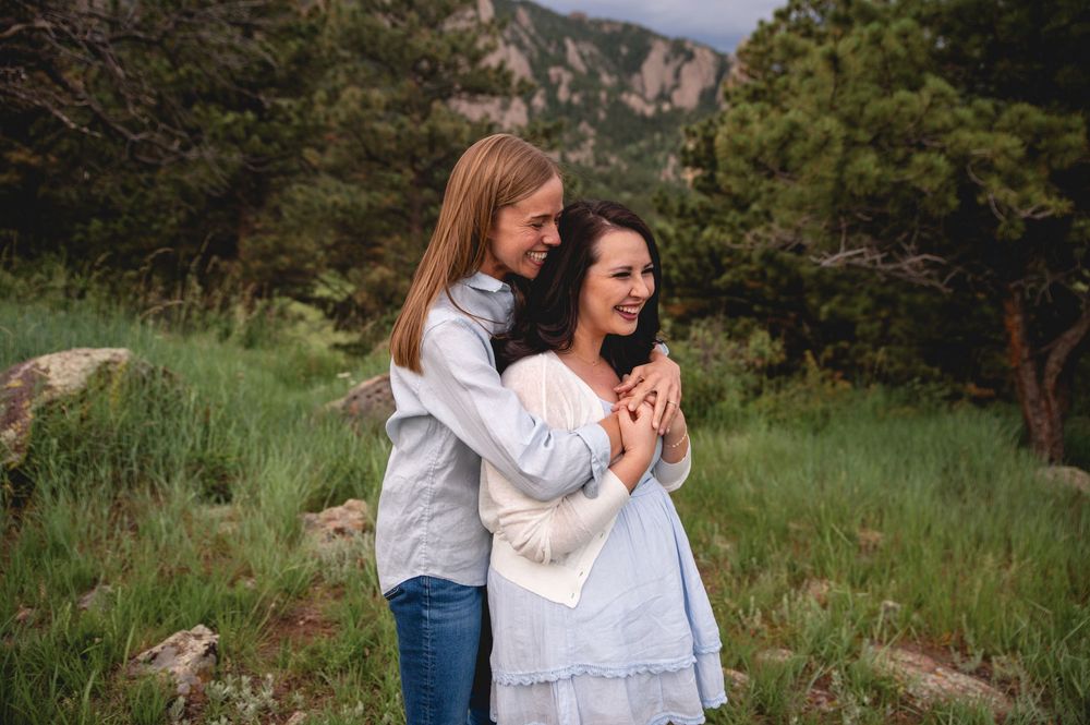 Two people in white tops share joyful moments together in a scenic mountain meadow with evergreen trees in the background.