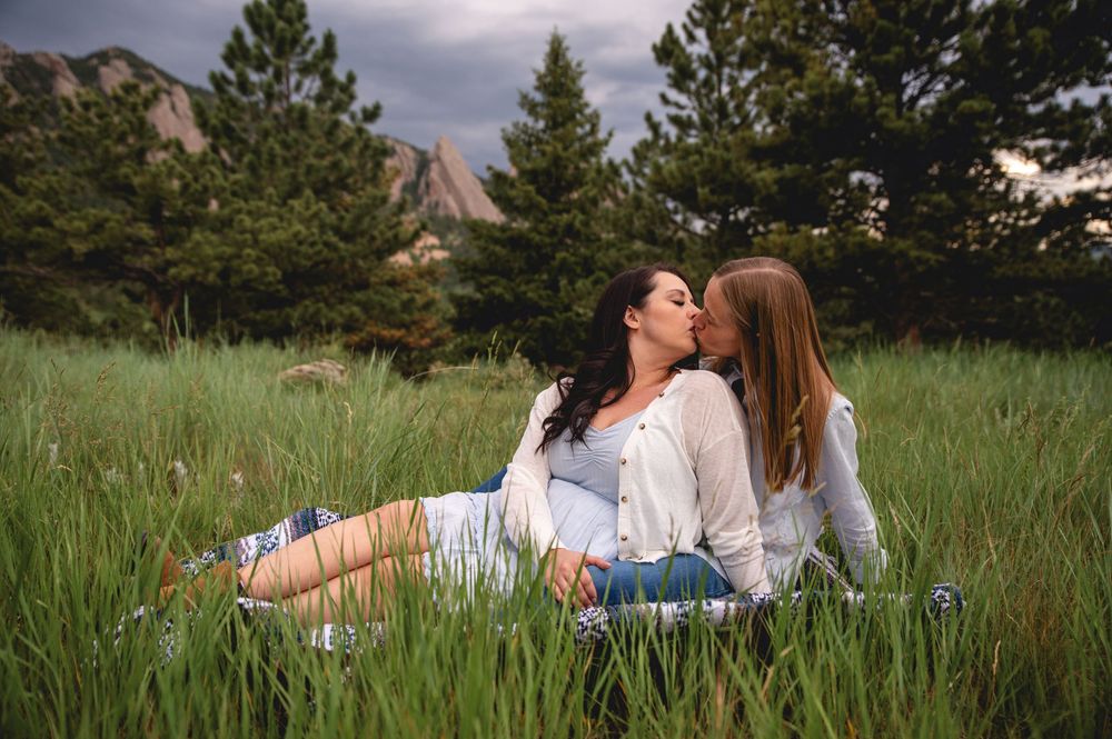 Couple lies together in a scenic mountain meadow surrounded by tall grass and pine trees during golden hour.