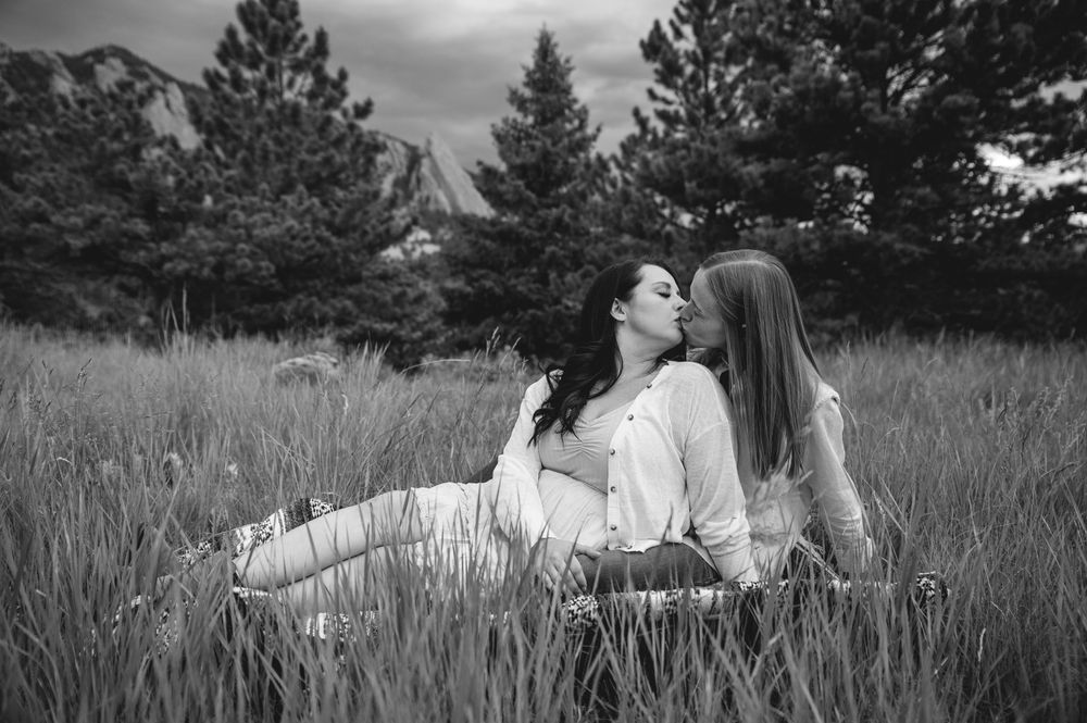 A romantic couple sits together in a meadow of tall grass with evergreen trees and mountains in the background.