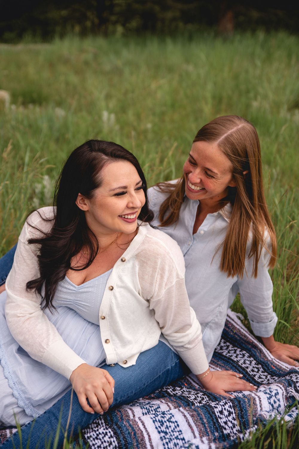Two friends share a joyful moment while sitting on a patterned blanket in a grassy field on a sunny day.