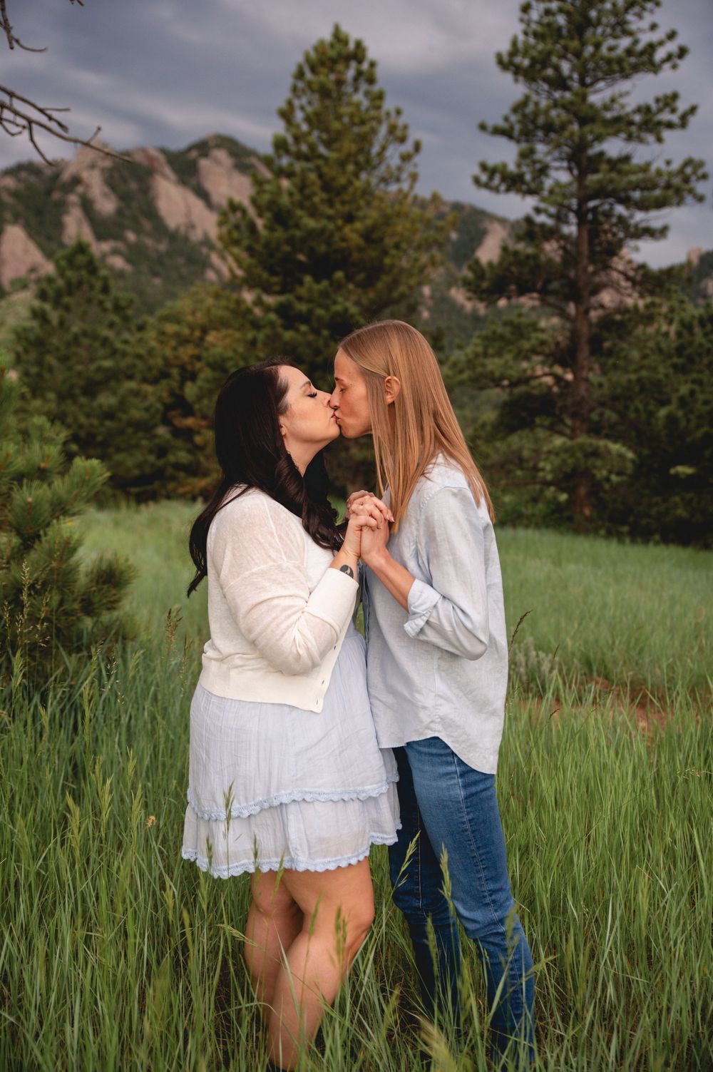 Romantic couple sharing a tender moment in tall grass with mountain landscape and pine trees in the background.