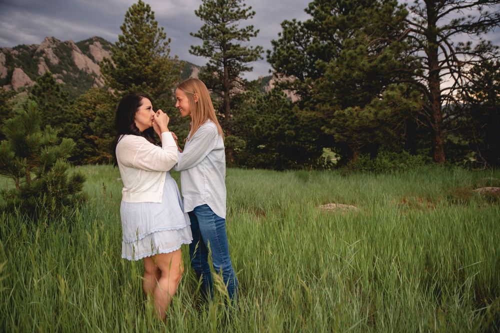 Two people share an intimate moment outdoors in a grassy field surrounded by tall trees and mountains at sunset.