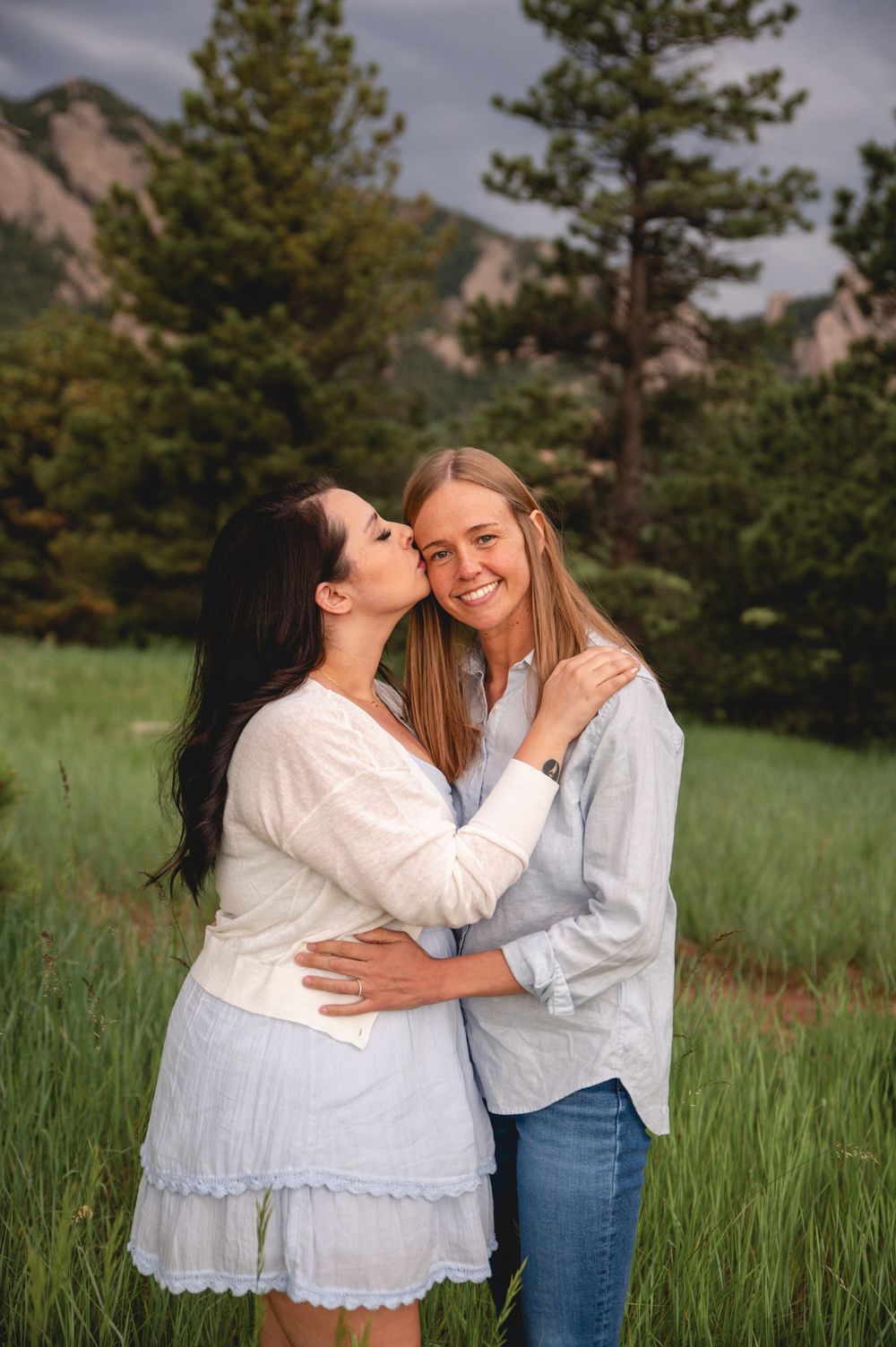 Two people embracing and laughing together in a field of tall grass with pine trees and mountains in the background.