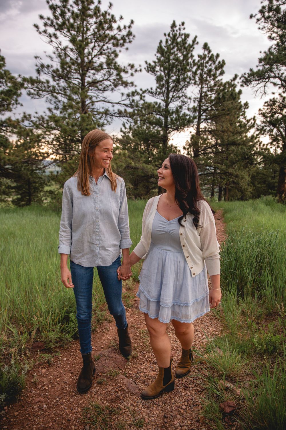 Two friends walking and chatting together on a dirt trail surrounded by pine trees and tall grass at sunset.