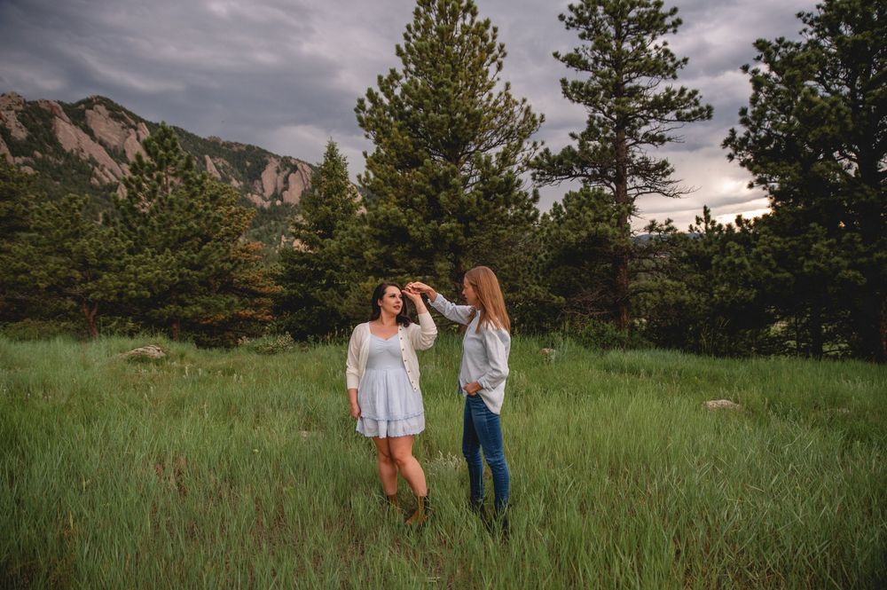 Couple walking through a green meadow with mountains and pine trees in the background under dramatic skies.