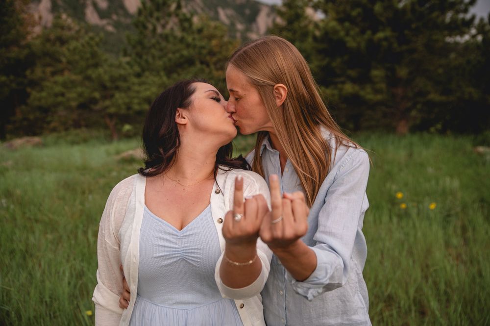 A couple in light colored outfits embrace and kiss in a scenic outdoor meadow setting with trees in the background.