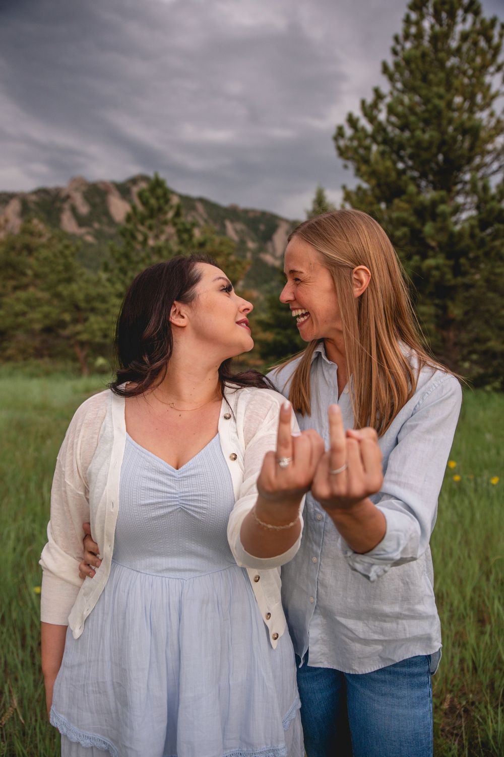Two friends sharing a playful moment while posing in a grassy field with mountains and evergreen trees in the background.