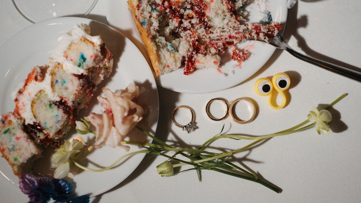 Flat lay composition with dessert crumbs, gold rings, googly eyes decoration and green plant stems on white surface.