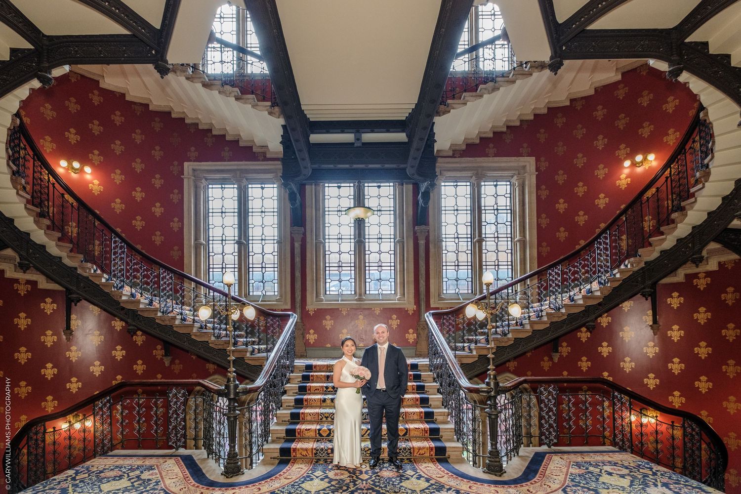 Elegant double curved staircase with red floral wallpaper and ornate railings inside a historic mansion.