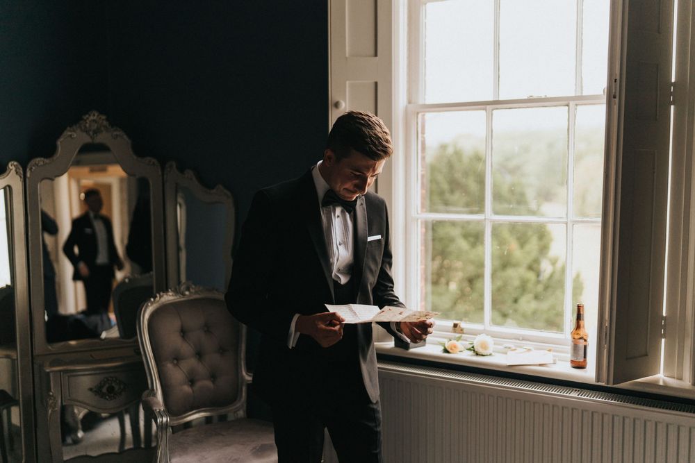 A groom in a black suit reads papers while standing by a bright window in an elegant vintage-style room at Hirst Priory.