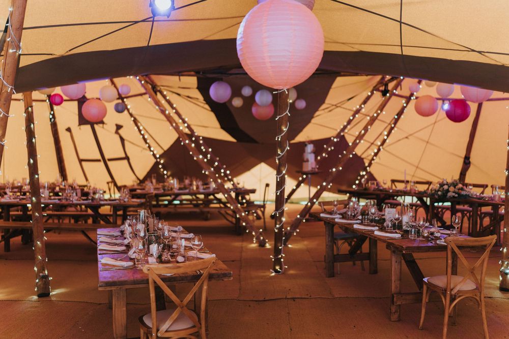Pink paper lanterns and string lights illuminate a rustic wedding reception setup inside a large tent venue at Hirst Priory.