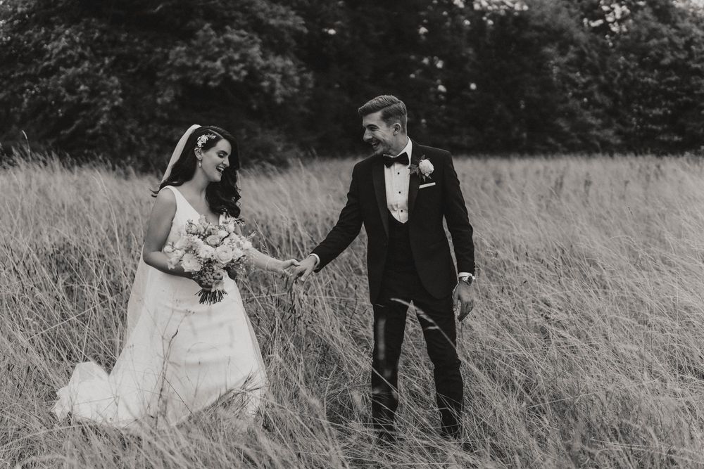 A wedding couple holds hands while walking through a tall grass field in black and white a Hirst Priory - taken by Grimsby photographers.