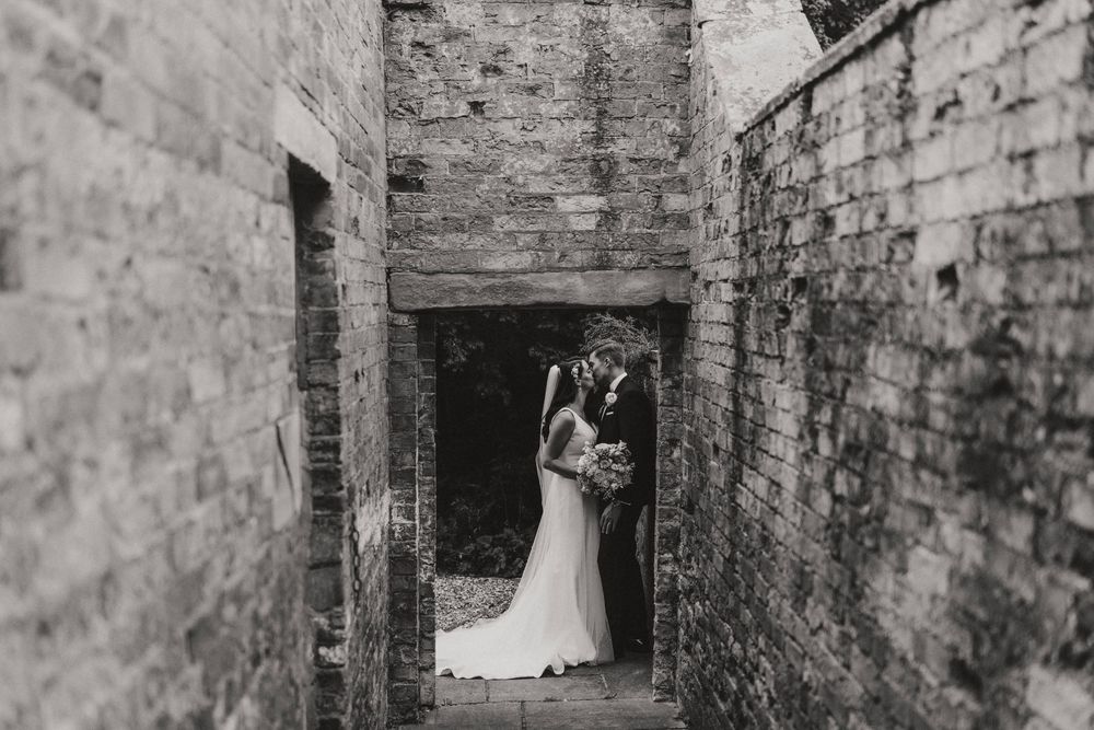A black and white photo of newlyweds embracing in an old brick alleyway or passageway at Hirst Priory.