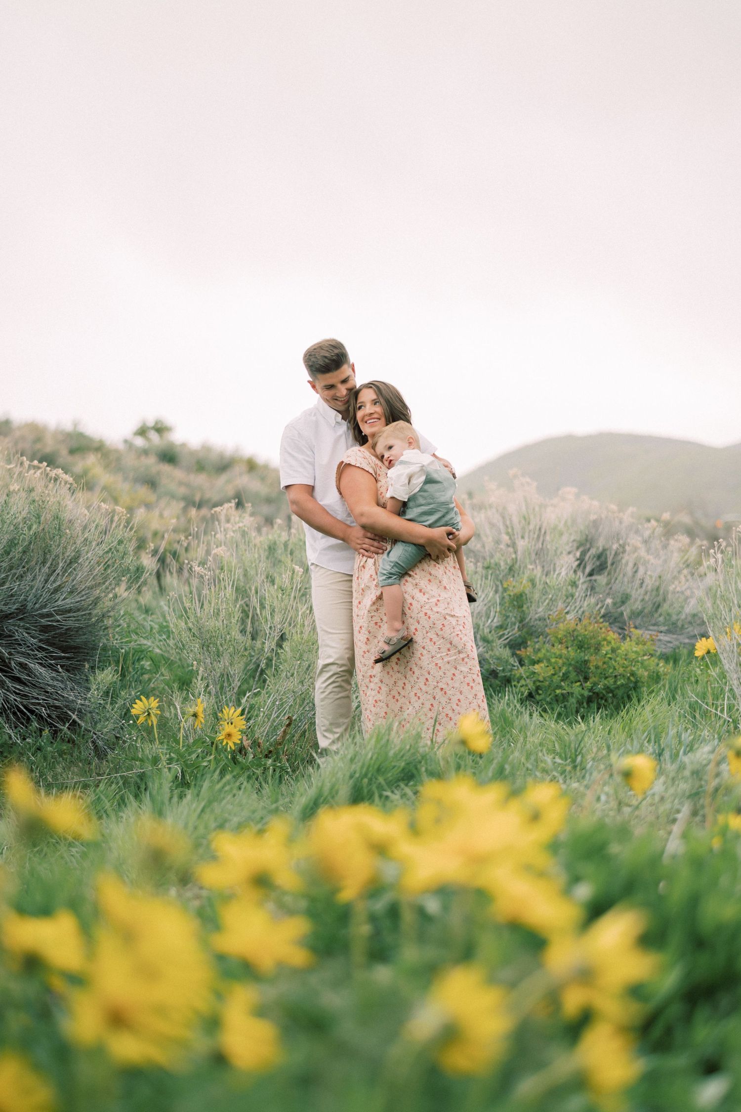 A couple embraces on a hilltop meadow filled with blooming yellow wildflowers under a soft pastel sky.