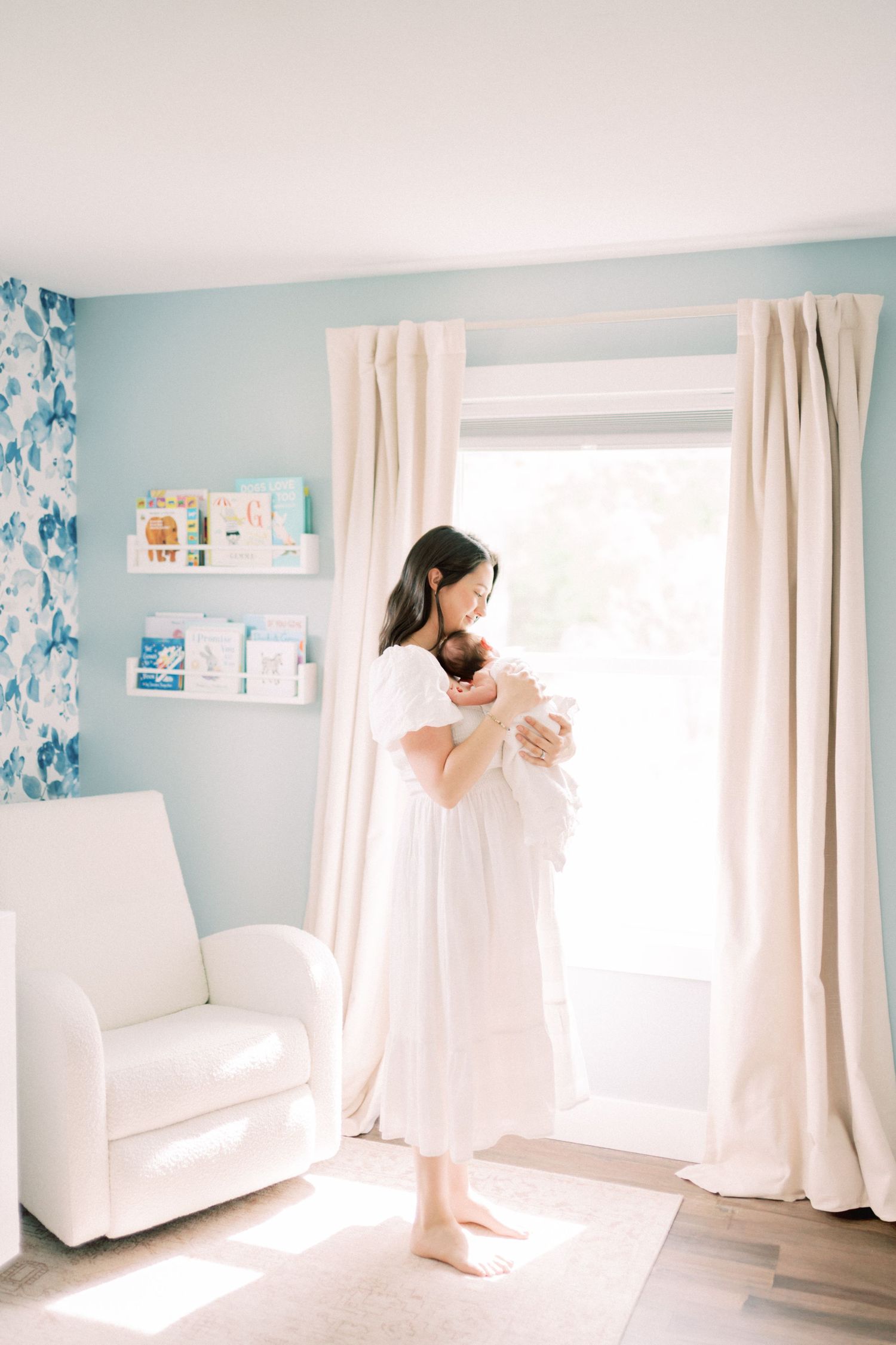 A sun-filled nursery with pale blue walls features a white armchair, floral wallpaper, and natural lighting through curtained windows.