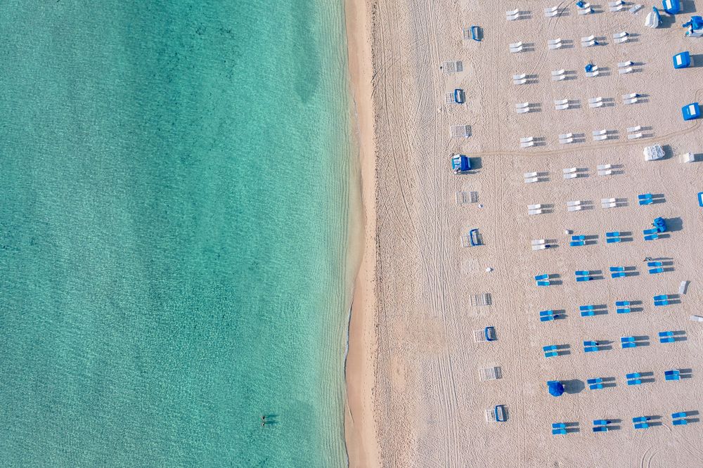 Aerial view of a pristine beach with turquoise water and rows of white and blue lounge chairs neatly arranged along the sandy shore.