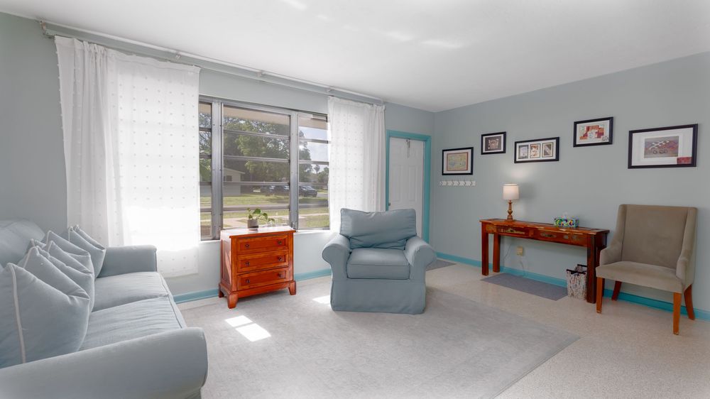 Sunlit sitting room with soft blue furniture, large window with sheer curtains, home office corner, and light neutral walls creating a calm,