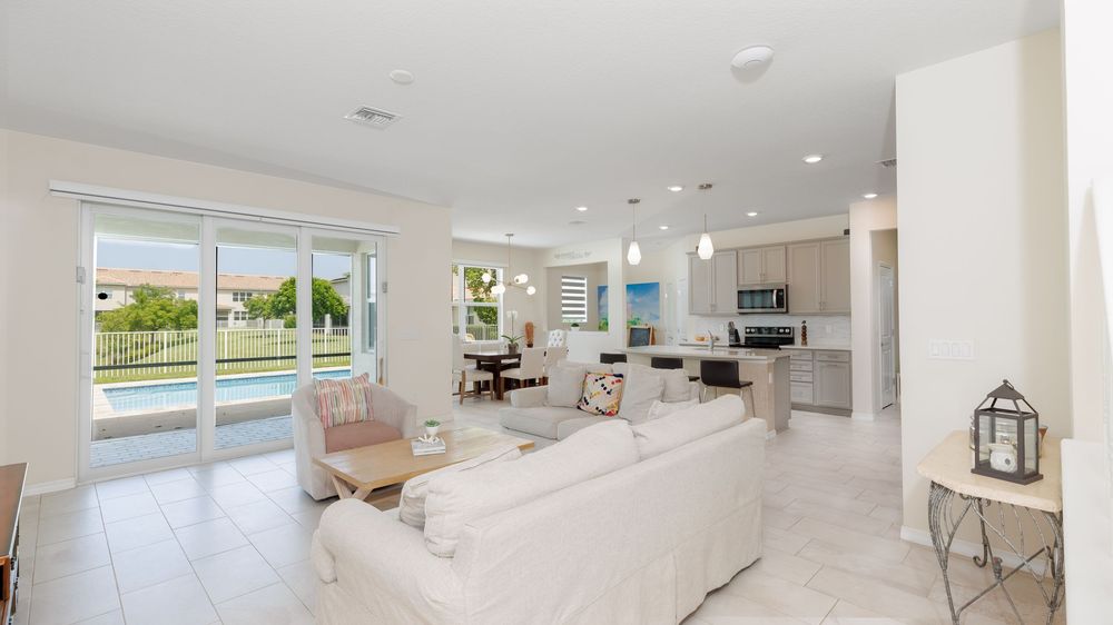 Bright open-concept living and kitchen area with neutral tones, tiled flooring, and large glass doors offering scenic views of a backyard po