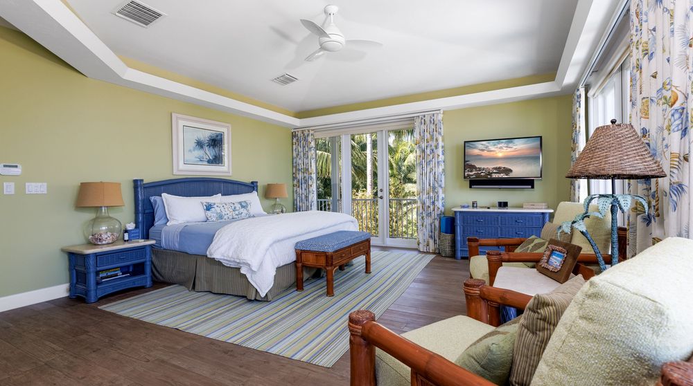 Tropical-themed master bedroom with blue coastal furniture, green accent walls, striped area rug, and French doors opening to a lush outdoor