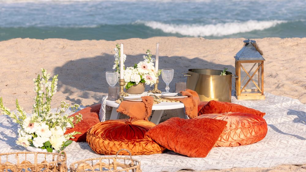 Boho-style beach picnic setup with decorative pillows, floral arrangements, elegant tableware, and ocean waves in the background.