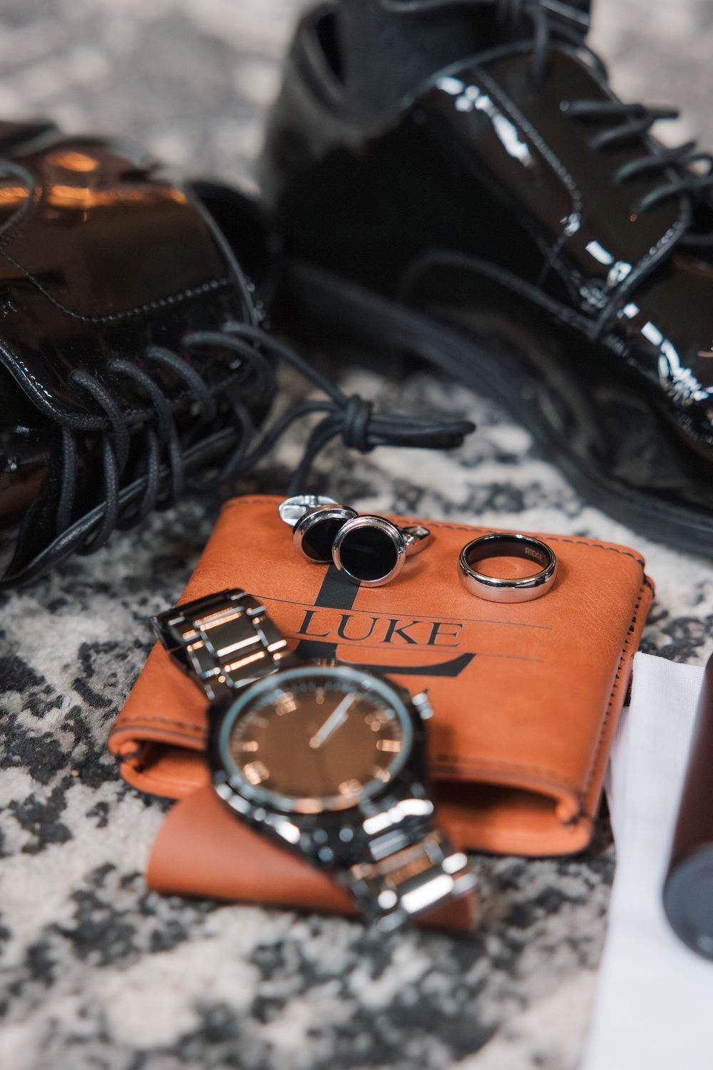 A stylish arrangement of black dress shoes, brown leather wallet, silver watch and men's accessories on marble surface.