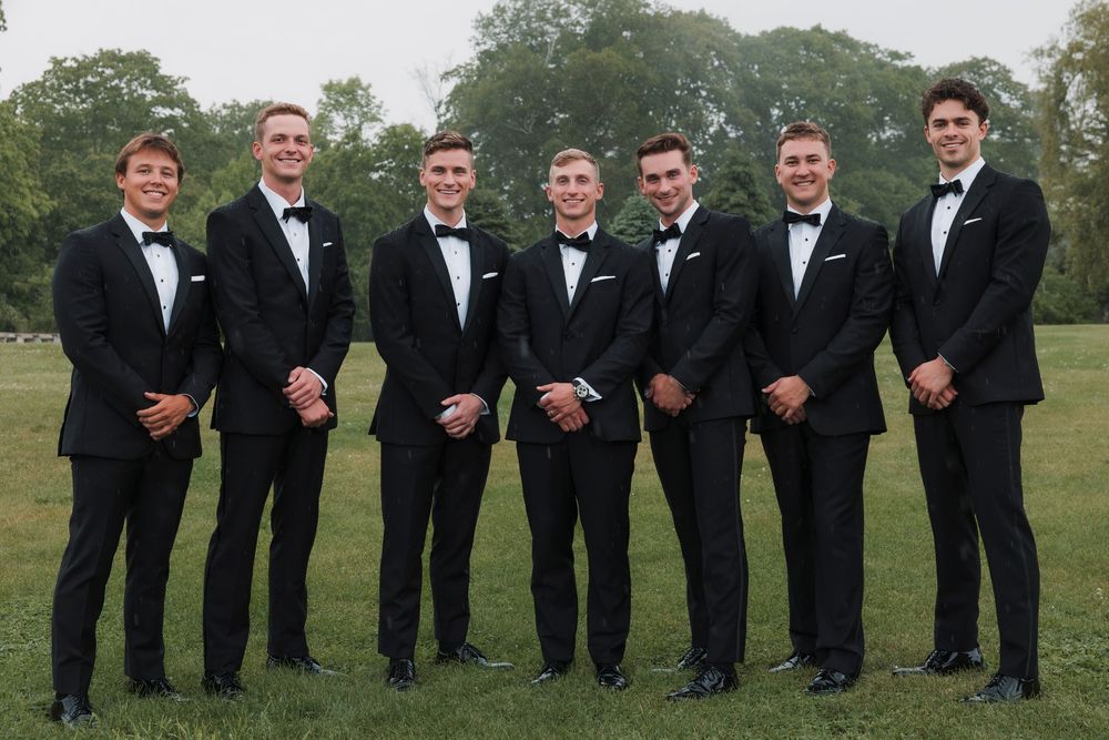 A lineup of groomsmen in classic black tuxedos and bow ties pose together outdoors.