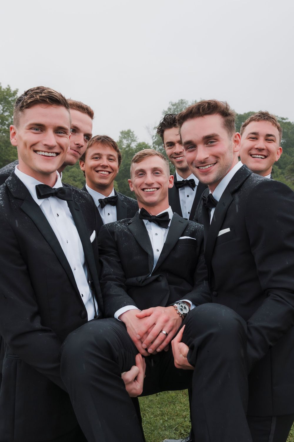Groomsmen in black tuxedos pose together sharing candid laughs and smiles during outdoor wedding photo session.