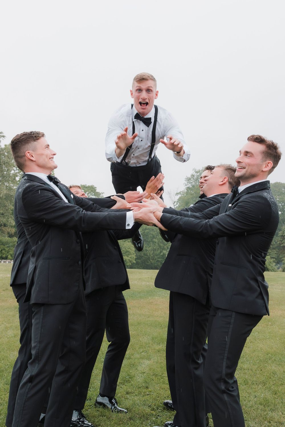 Groomsmen in black suits playfully toss their celebrating companion in the air on a grassy lawn.