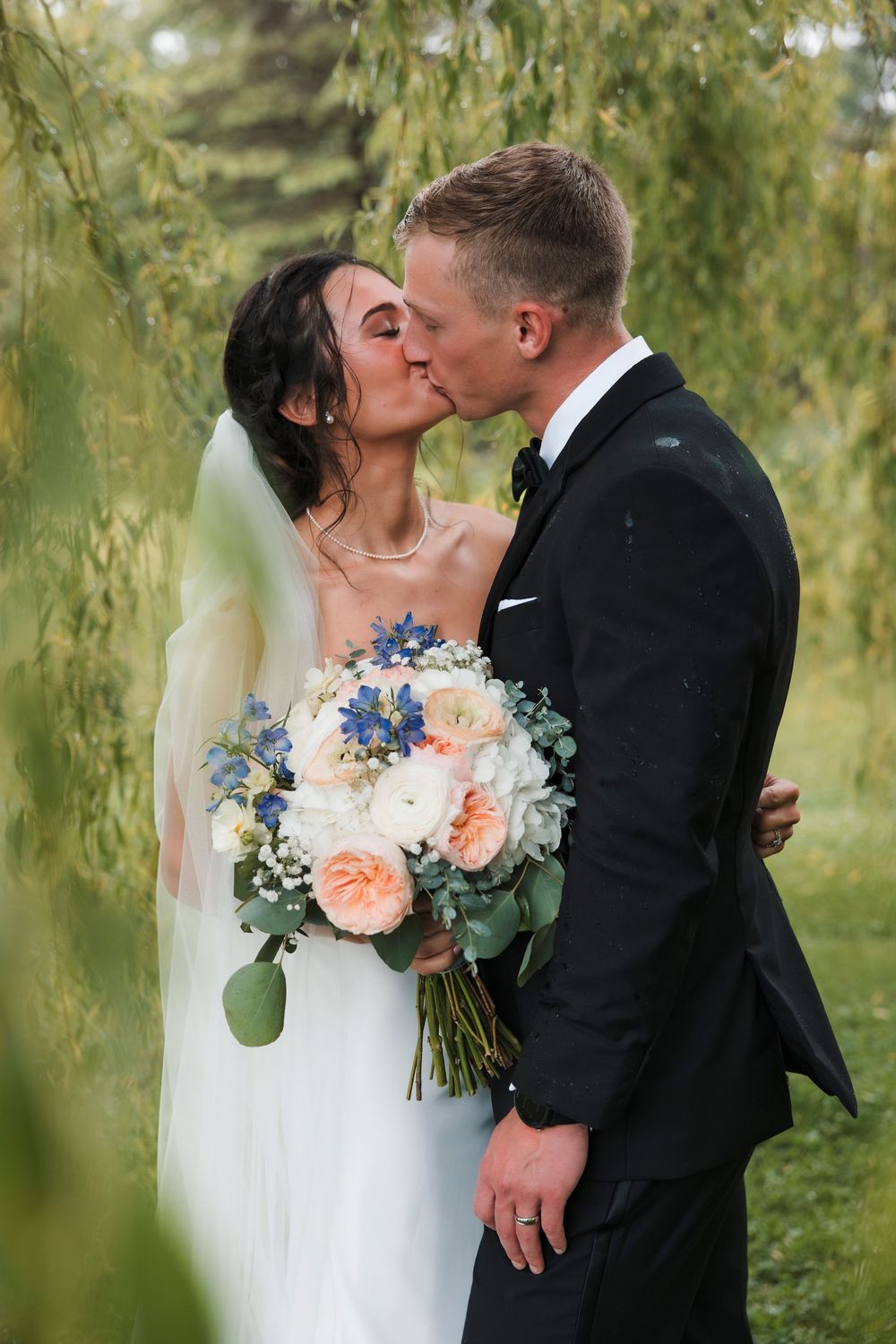 A couple shares romantic kisses in an outdoor garden setting with a bridal bouquet featuring blue and peach flowers.