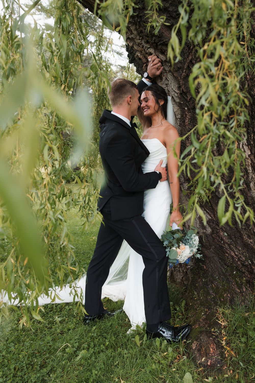 A bride in white dress and groom in black suit share romantic moments under a weeping willow tree during their photoshoot.
