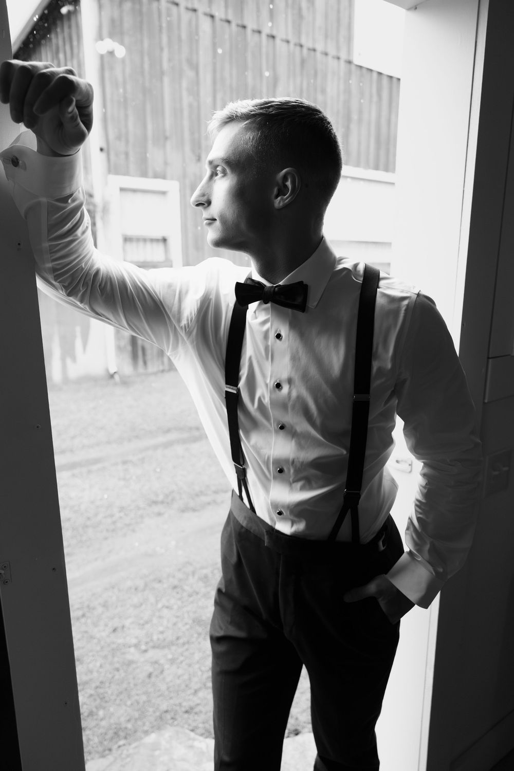 Black and white portrait of a formally dressed person in suspenders and bow tie leaning against a doorframe.