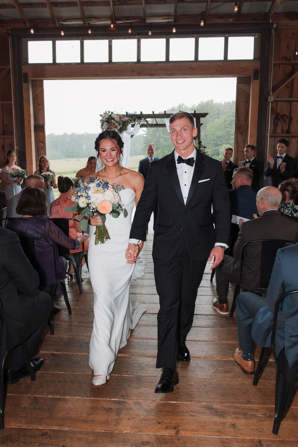 Newlyweds walk down rustic wooden aisle together after ceremony in barn venue with natural light streaming through windows.