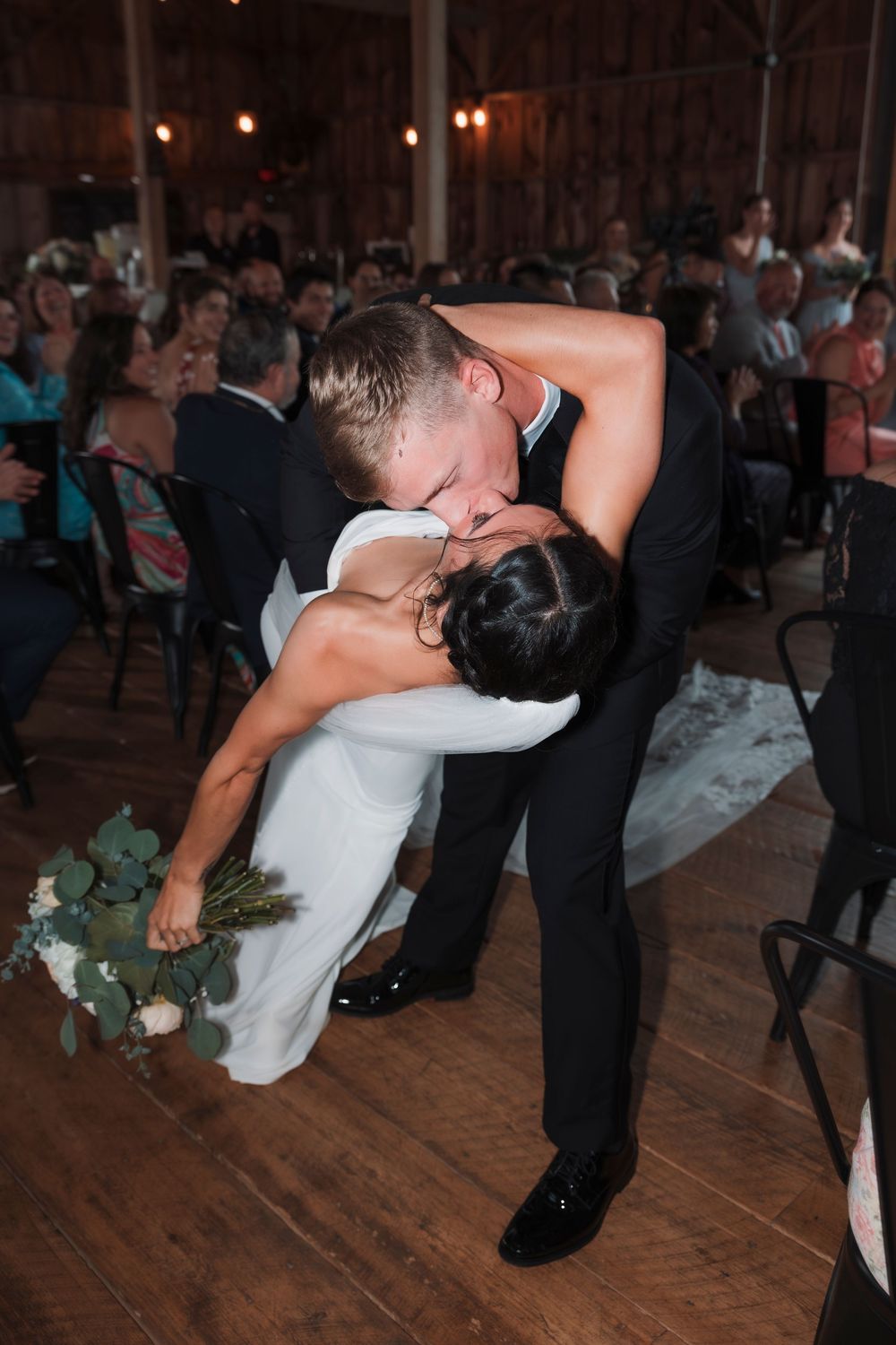 A romantic dip during a first dance at a reception, with guests watching on a rustic wooden dance floor.