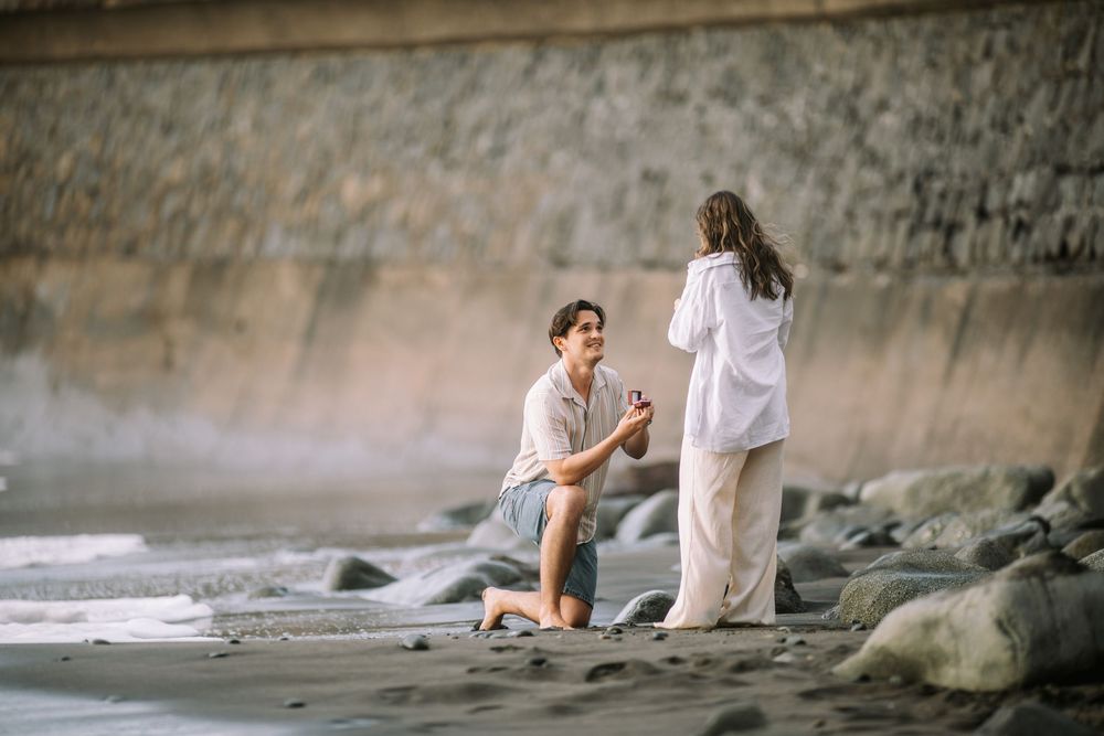 A romantic beach proposal scene with rocky shoreline and concrete wall in the background.