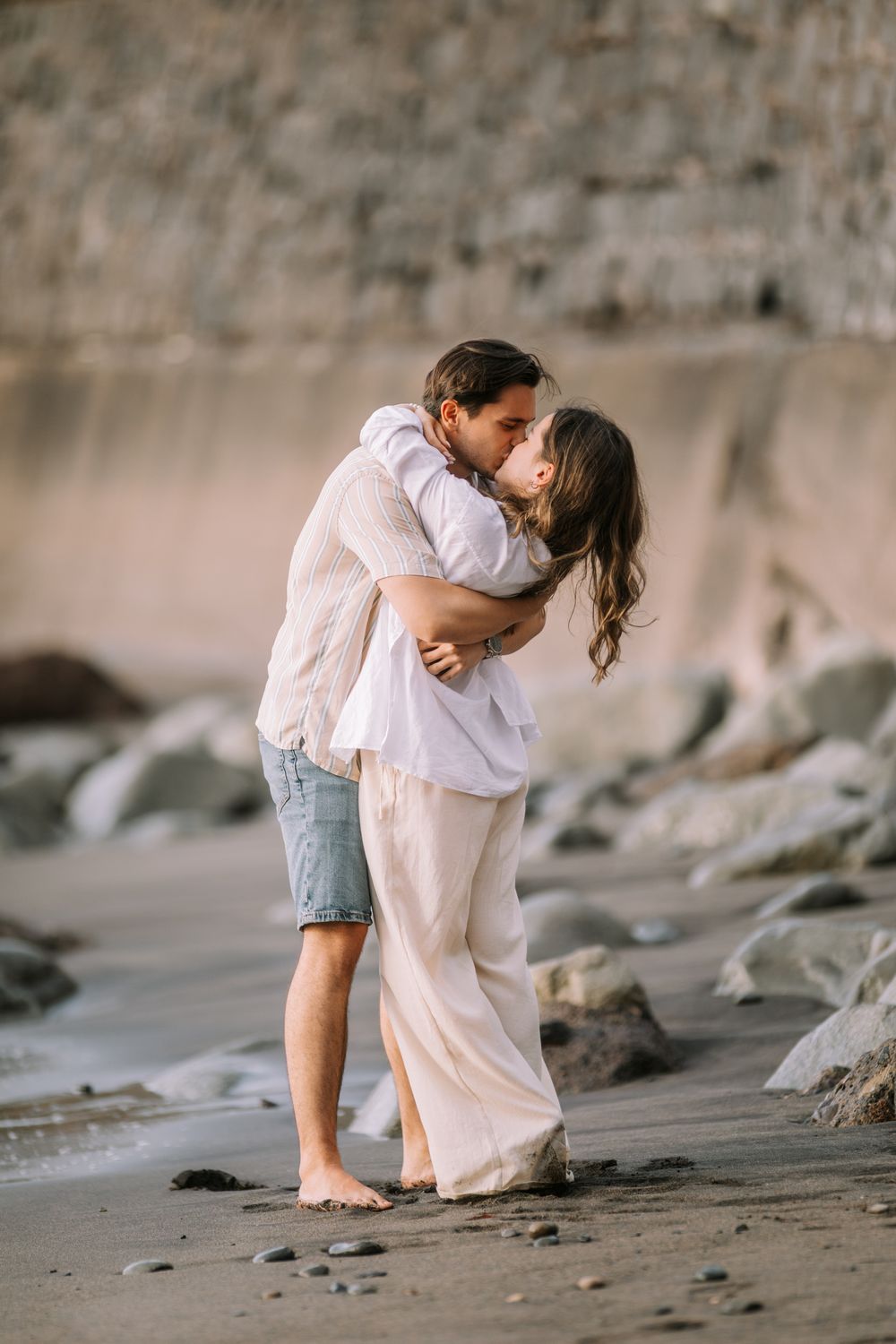 A couple shares an intimate romantic moment on a sandy beach at dusk with cliffs in the background.