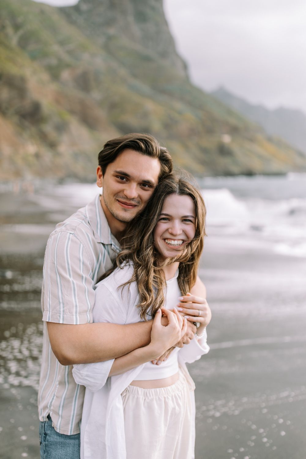 A happy couple embraces and smiles together on a scenic beach with dramatic coastal cliffs in the background.