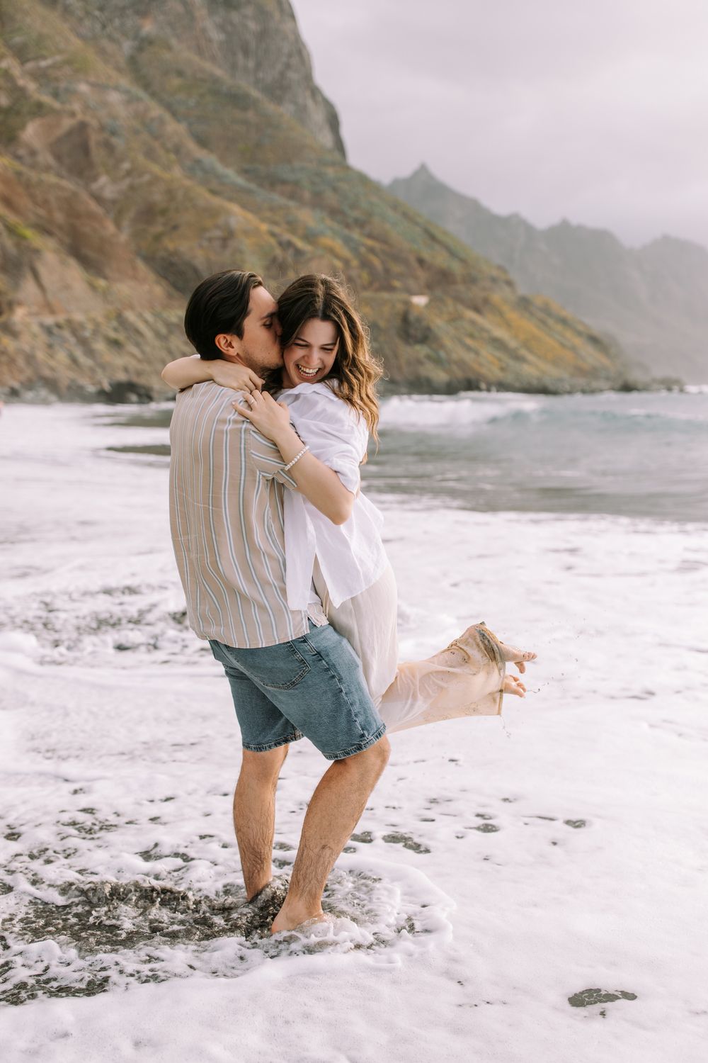 A romantic couple embraces on a misty beach with dramatic coastal cliffs in the background.