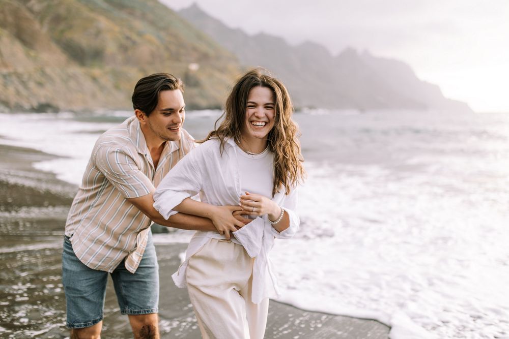 Happy couple laughing and running along a misty beach coastline at sunset.