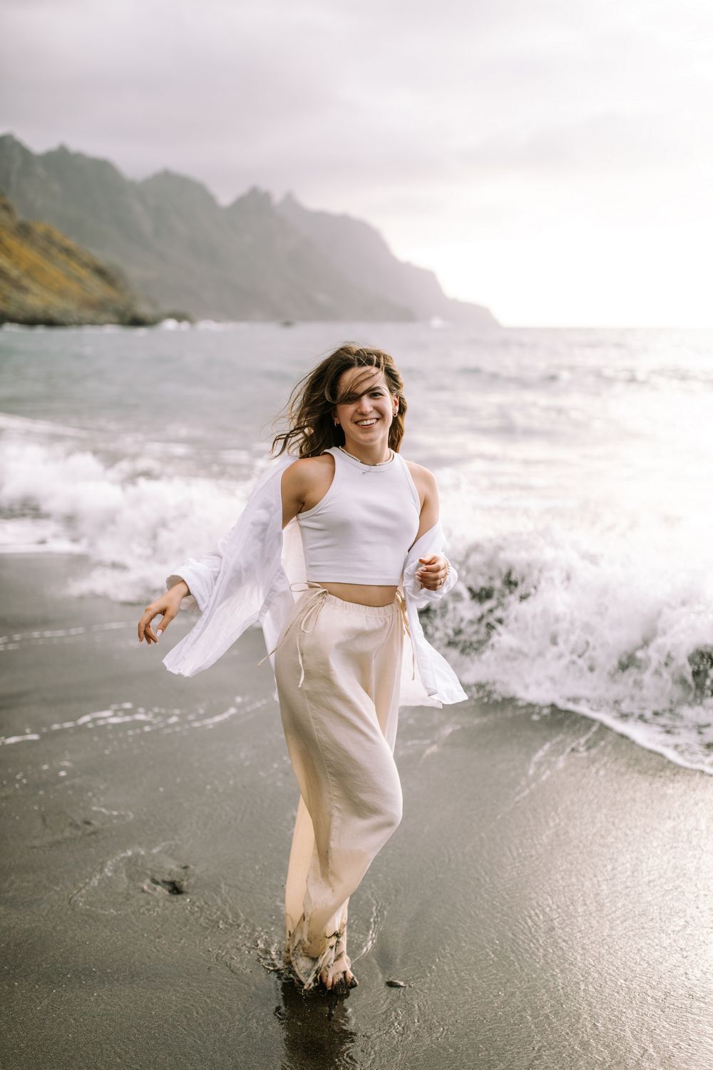 Someone in white flowing clothing stands joyfully on a misty beach with dramatic mountains in the background.