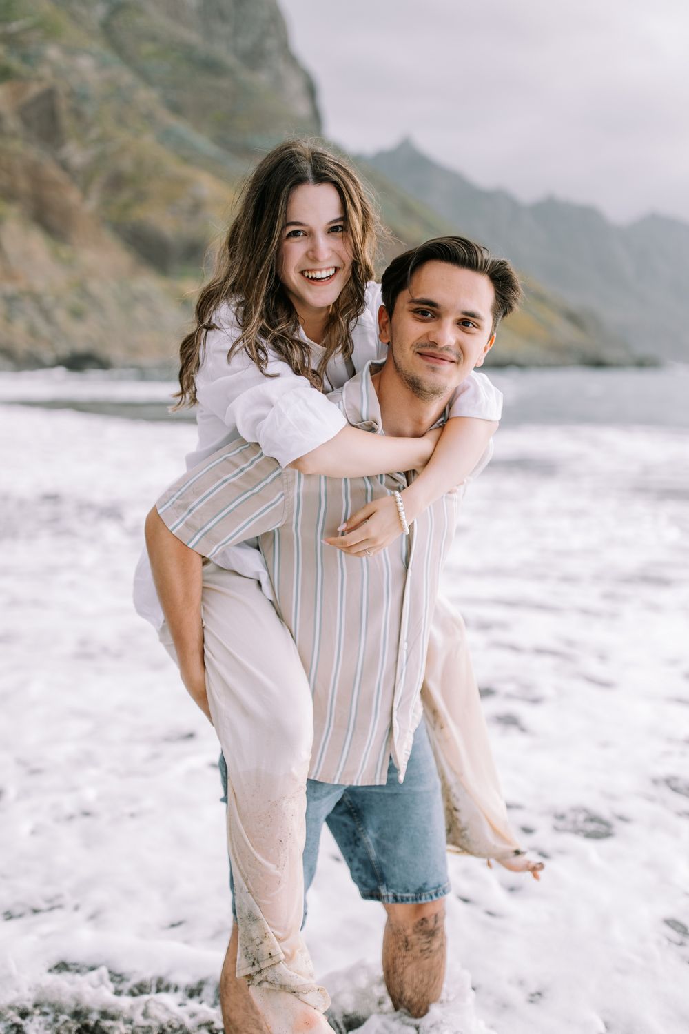 A couple poses together on a scenic beach with mountains in the background on a cloudy day.
