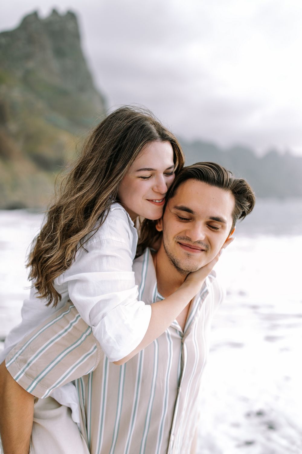Couple embracing on a misty beach with dramatic coastal cliffs in the background.