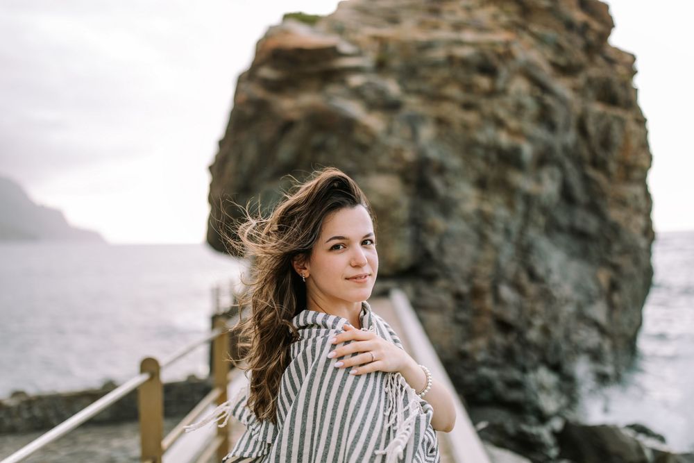 Person in striped blouse poses on rocky coastal cliffs as wind blows through their hair during sunset.