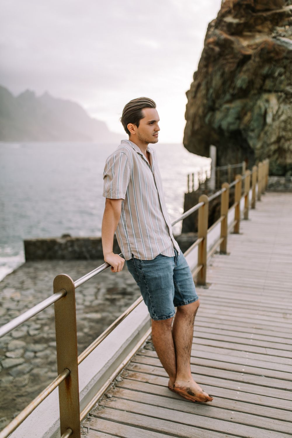Person in striped shirt and denim shorts stands barefoot on wooden boardwalk along misty rocky coastline.