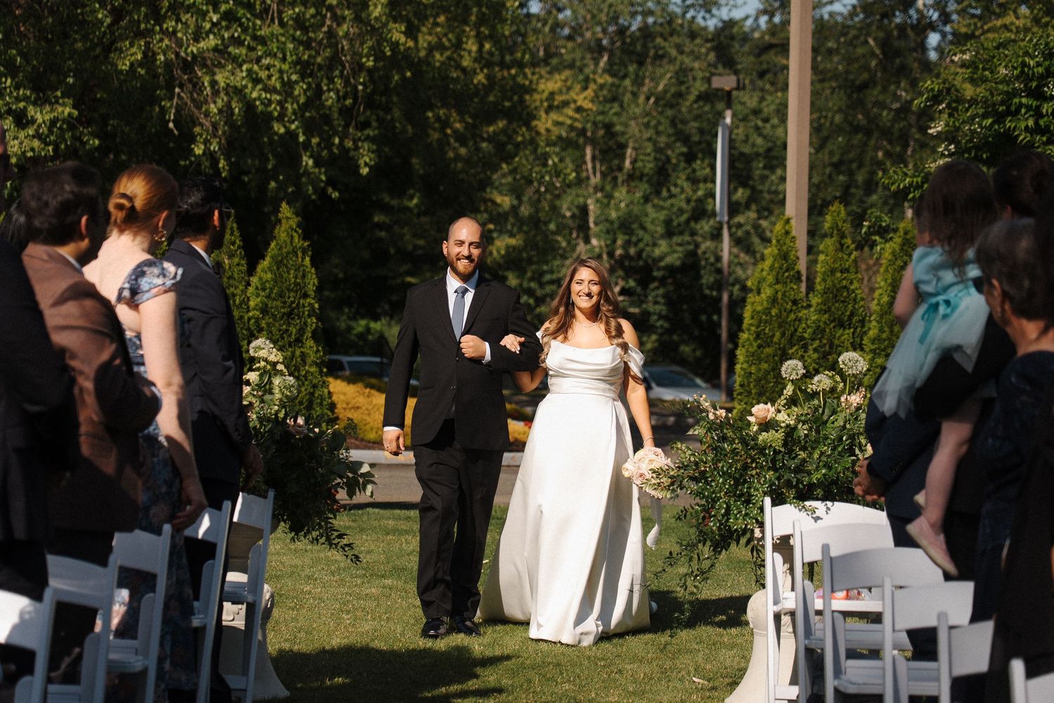 A couple walks down an outdoor garden aisle after their wedding ceremony on a sunny day.