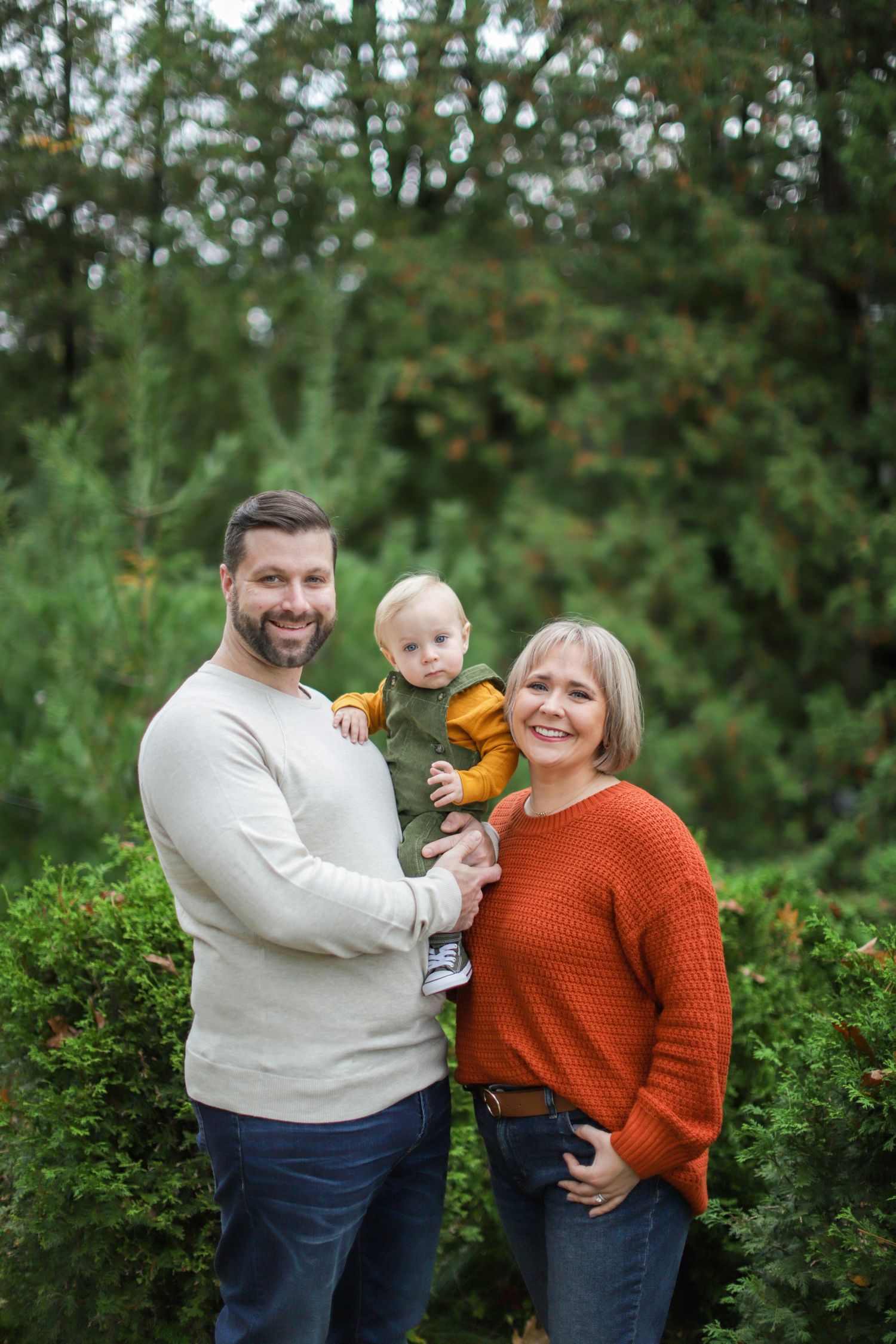 Three people in casual clothing laugh together while sitting on a forest slope surrounded by tall pine trees.