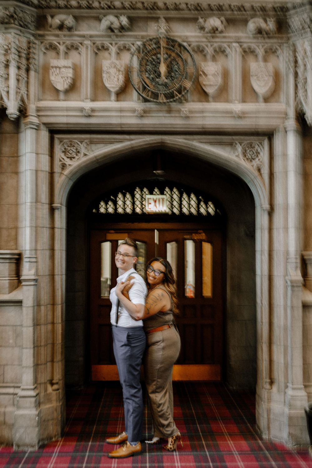 A couple embraces in a romantic pose under an ornate Gothic archway with decorative stonework and a plaid carpet.