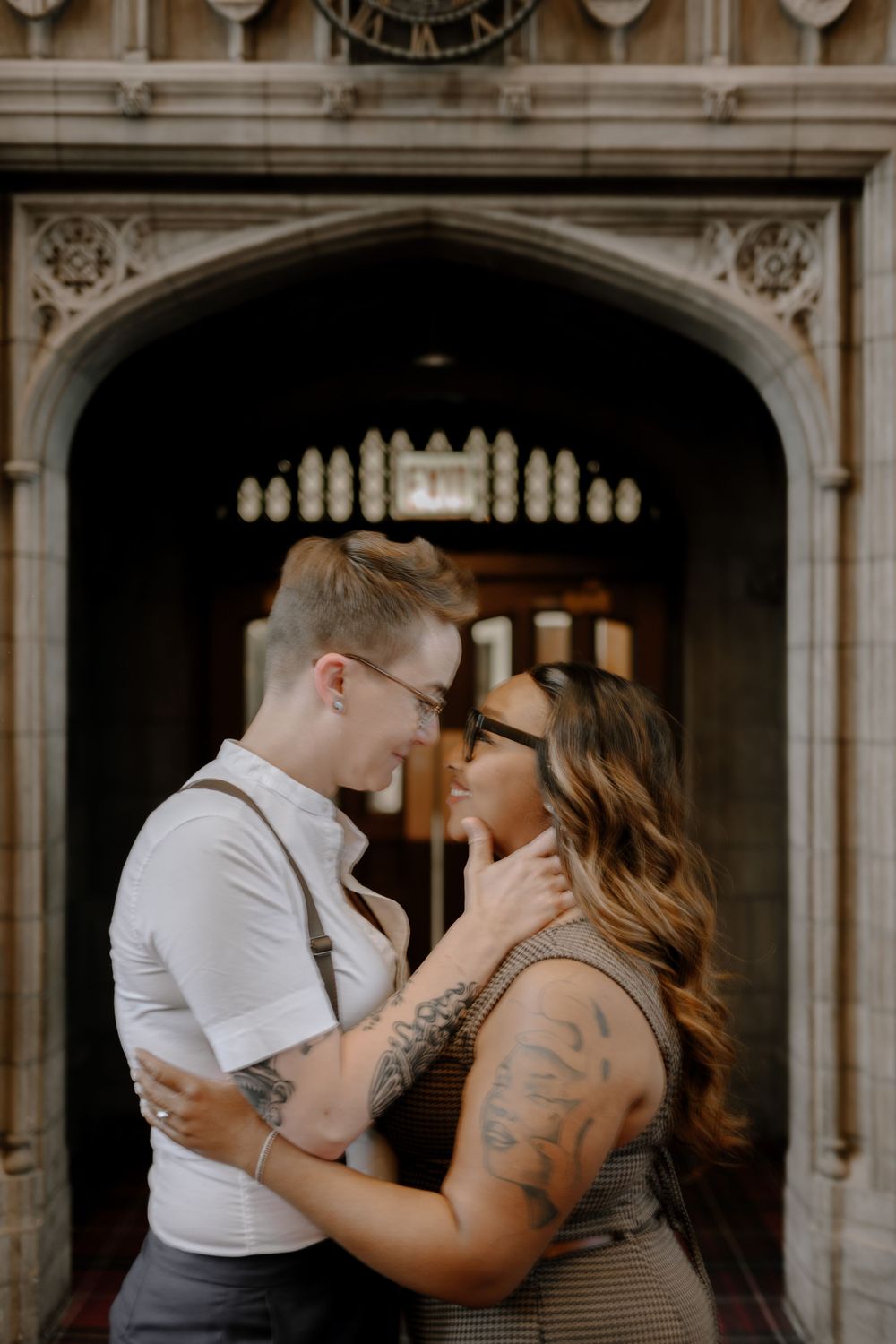 A romantic couple embraces in front of an ornate stone archway with decorative gothic architecture.