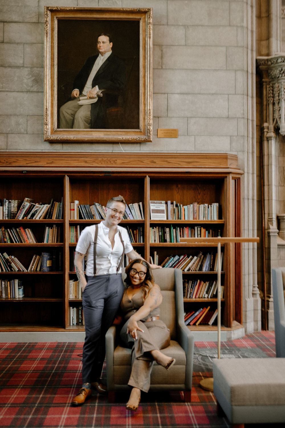 A stylish couple posing in front of a library bookshelf with an old portrait painting hanging above.